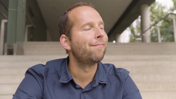 A Caucasian Man Looks Around with a Smile As He Sits on a Staircase in an Urban Area