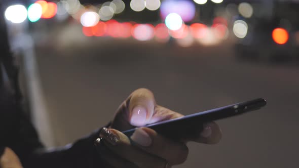 Close Up Woman Using Smartphone in the City at Night Time. Girl Looks at the Smartphone Screen alt