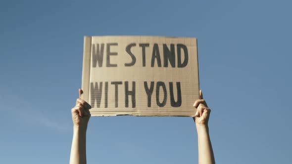 Sign WE STAND WITH YOU against blue sky. Woman hands waving poster "We Stand With You". alt