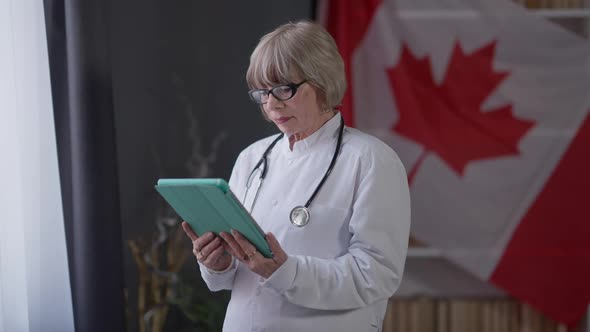 Concentrated Senior Female Doctor Reading Latest News Online Standing with Tablet Indoors alt