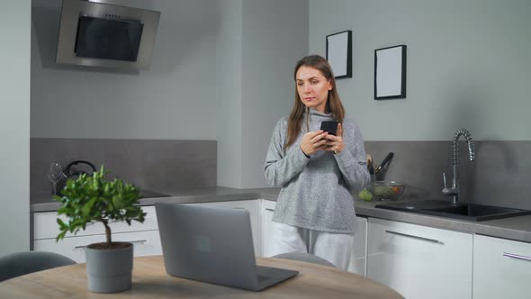 Woman with Smartphone in Hand Standing in Home Cozy Kitchen Use Laptop Do Freelance Work Looking alt