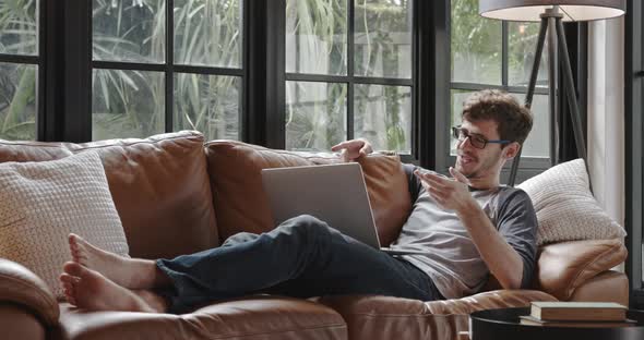 Young happy Caucasian man chatting online via video call with laptop computer on the couch at home