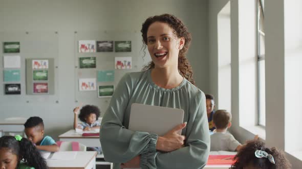 Video of happy caucasian female teacher holding laptop in class of diverse pupils alt