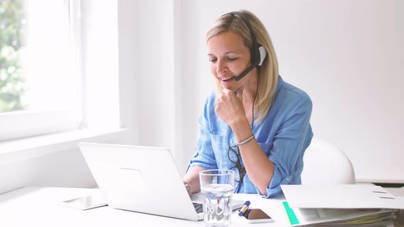 Businesswoman sitting at desk using headset and laptop alt