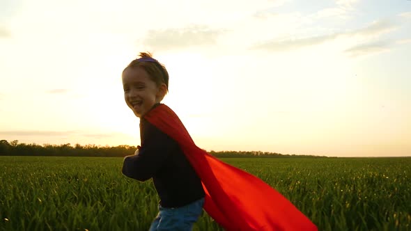 Cheerful Boy Running on a Green Meadow at Sunset. Happy Kid in Superman Costume alt