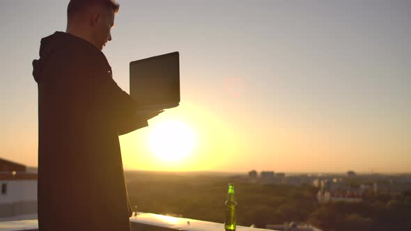 A Programmer with a Laptop Sits on the Roof and Writes Code at Sunset. Remote Work Freelancer alt