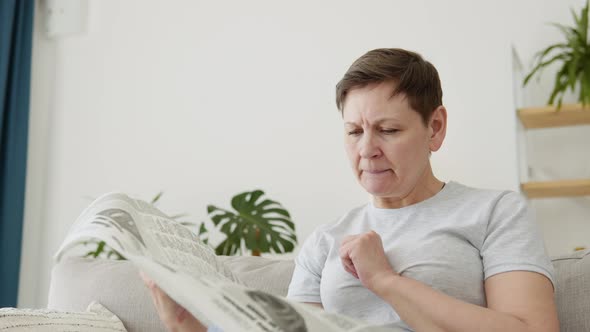 A Closeup View of a Beautiful Mature Woman is Reading a Newspaper Sitting on Sofa at Home in the alt