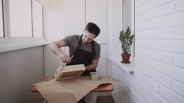 Man in Black Apron Covers Surface of Wooden Box with Colorless Varnish alt