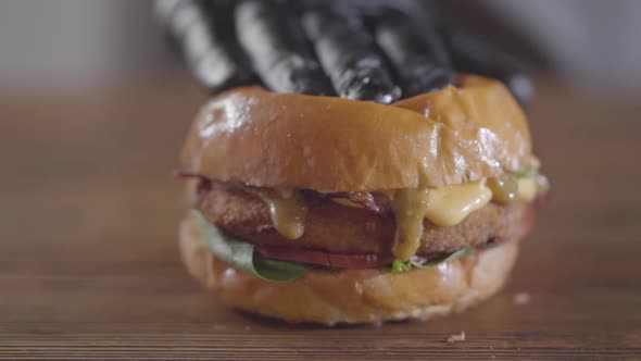 Hand of the Chef in Black Rubber Gloves Pressing Burger and Sauce Flows Out alt