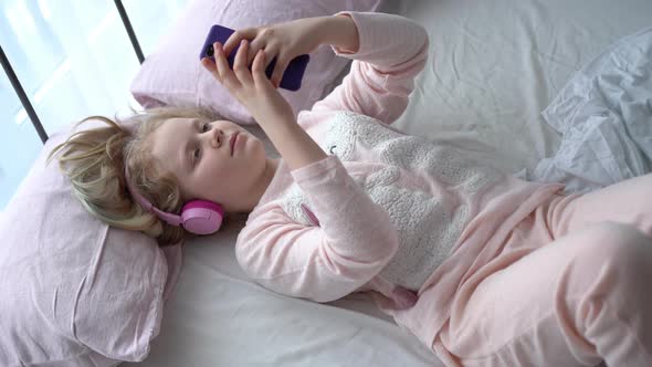 Modern Life of Generation Z. Teenage Girl in Pajamas and Headphones in the Room on the Bed Listens