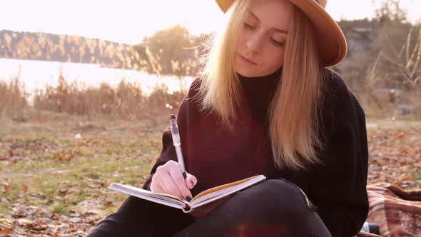 Slow Motion Caucasian Blonde Woman with Beige Hat in Black Sweater Write on Notebook in the alt
