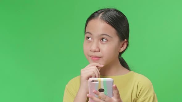 Kid Girl Use Mobile Phone And Thinking About Something While Standing On Green Screen In The Studio alt