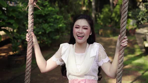 slow-motion of happy young woman in Thai traditional dress relaxing on a wooden swing alt
