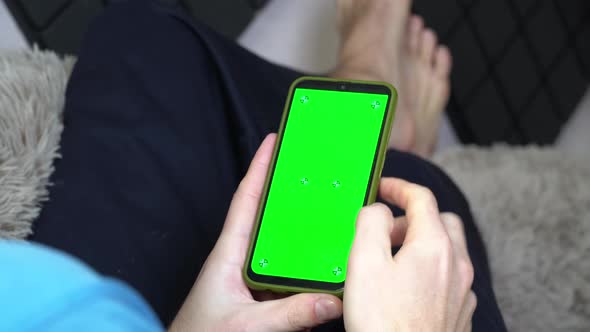 A Man on the Couch With a Phone in His Hands With a Green Screen Back View alt