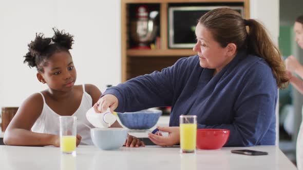 Happy caucasian lesbian couple and their african american daughter eating breakfast in kitchen alt