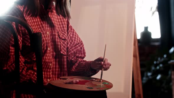 A Young Pretty Woman Artist Mixing Warm Colors on the Palette Sitting in Front of an Easel alt