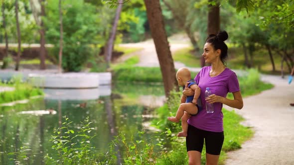 Fit Woman Carrying Baby and Drinking Water After Training in Park alt