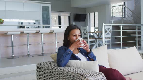 Mixed race woman sitting on couch drinking cup of coffee alt