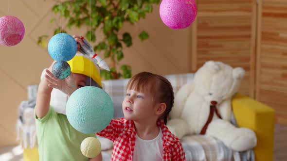 Boy and a Girl Play Astronauts Flying a Toy Rocket Among Planets on Strings in Children's Room alt
