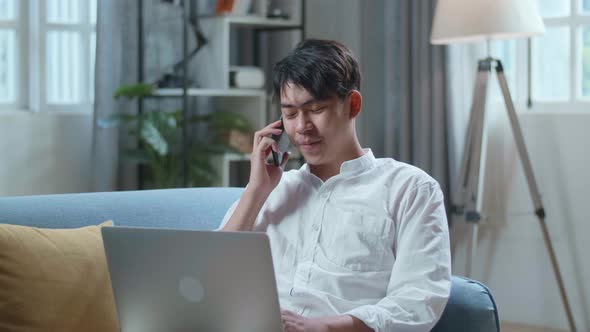 Smiling Asian Man Answering The Call On Smartphone While Lying On Sofa And Using Laptop Computer alt