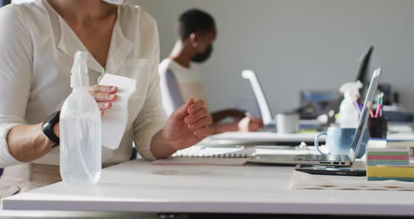 Two diverse female colleagues wearing face mask, sanitizing desk in office alt