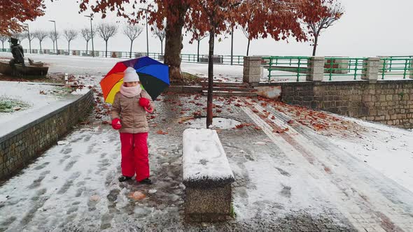 Little child girl with umbrella of many colors plays kicking snowball. Slow-motion alt