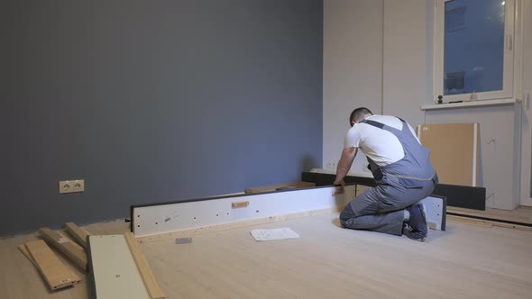 A Builder in a Work Overalls is Assembling a Bed in a New Apartment alt