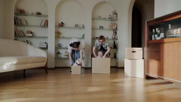 Excited Happy Young Parents Run Push Cardboard Boxes with Little Cute Girl Daughters Ride Insideand alt