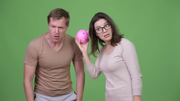 Young Couple Shaking Piggy Bank Together Against Green Background alt