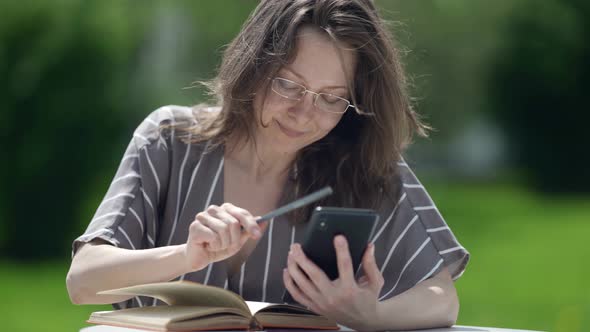 Pretty Adult Woman is Reading Book in Foreign Language and Using Smartphone for Translating alt