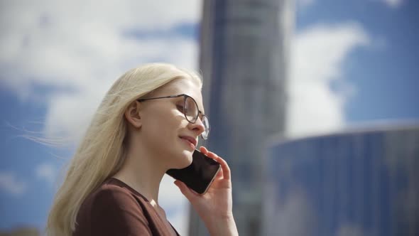 Young Blonde Woman in Glasses Talking on the Phone Against the Backdrop of a Glazed Building