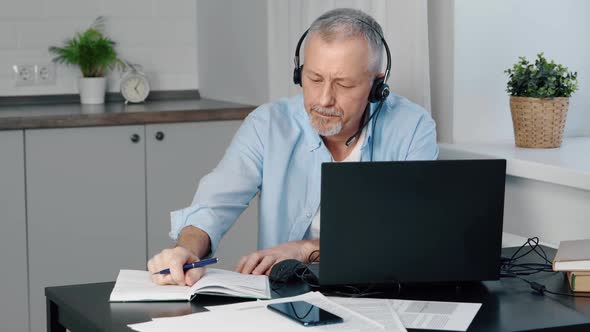 An Elderly Man in Headphones and a Computer Takes Notes in a Notebook