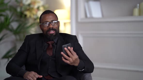 African-American with a Beard, Black Suit, Stylish Glasses. The Businessman Is Sitting on a Chair alt