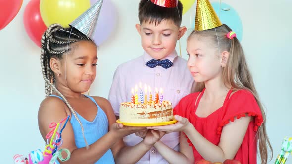 Little Girls Hold Cake While Boy Blows Candles at Birthday alt