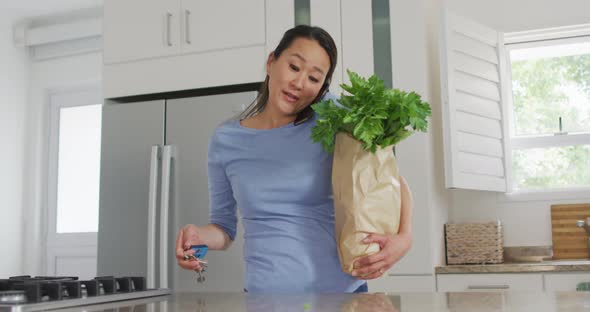 Asian woman coming back home with groceries and having call on smartphone alt