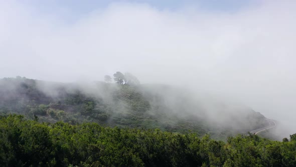 Flying Through the Clouds Over a Mountain Landscape and Road Surrounded By Green Vegetation alt