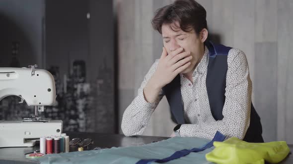 Handsome Brunette Man Yawning and Putting Head on the Table Next To Sewing Machine. Portrait  alt
