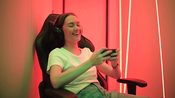 Cute Gamer Girl Sit on a Gaming Chair, Smiling and Playing Mobile Online Game on a Smartphone alt