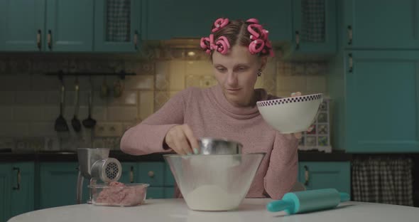 Funny Young Woman Kneading Dough Sitting in the Kitchen. Housewife with Hair Curlers on Head Mixing alt