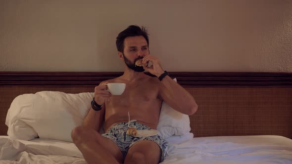 Man Eating Croissant In Bed.Man Lying And Taking Breakfast In Bed Enjoying Coffee. alt