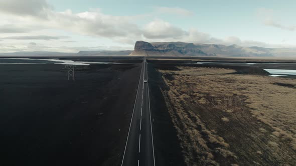 Car drives long straightaway through black sand desert in Iceland. Aerial follow shot. alt