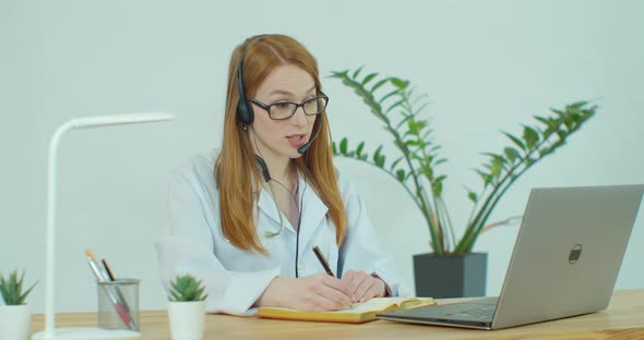 Female Medical Assistant Wears White Coat, Headset Video Calling Distant Patient on Laptop. Doctor alt