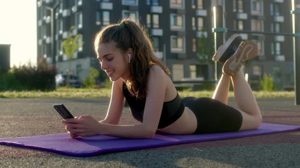 Cheerful Happy Young Woman Lies on Fitness Mat During Break Between Outdoor Sports and Communicates alt