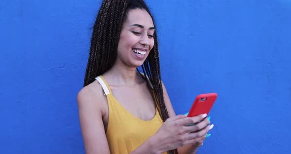 Bohemian mixed race young woman using smartphone outdoor with blue background alt