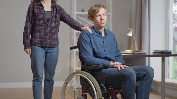 Sad Depressed Disabled Man in Wheelchair Thinking As Unrecognizable Woman Putting Hand on Shoulder alt