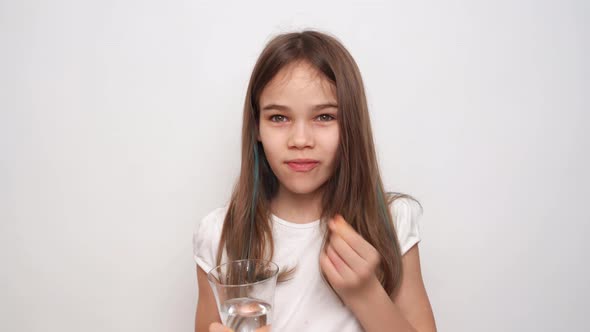 A Teen Girl Puts a Pill in Her Mouth and Wash Down with Water Against White Wall alt