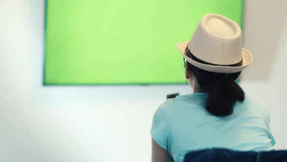 Brunette Girl in a Hat Sits in Font of a Geen Sreen TV Switches Channels Green Chroma Key Screen TV