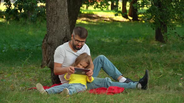 Happy Family Father and Girl Sit on Lawn in Summer Park with Digital Tablet