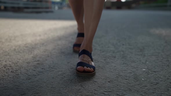 Female Feet in Black Sandals are Walking on the Asphalt alt
