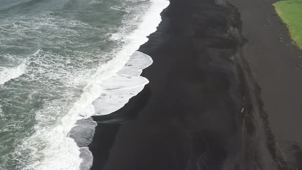 Black sand beach with waves in Vik, Iceland with drone video above moving back. alt